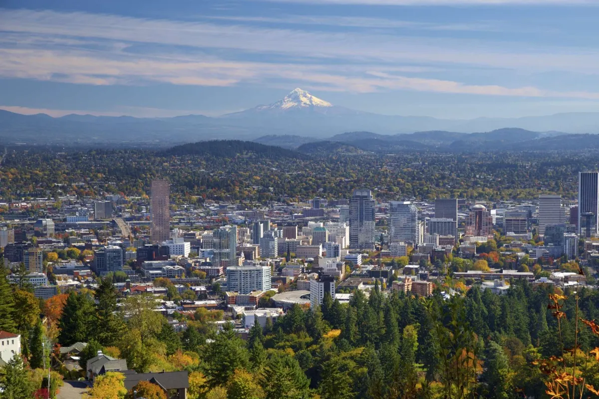 A stunning view of Portland's downtown skyline framed by vibrant autumn foliage, with the iconic Mount Hood rising majestically in the background. The cityscape glows in the golden hour, and the river reflects the colors of the sunset and snow-capped mountain.