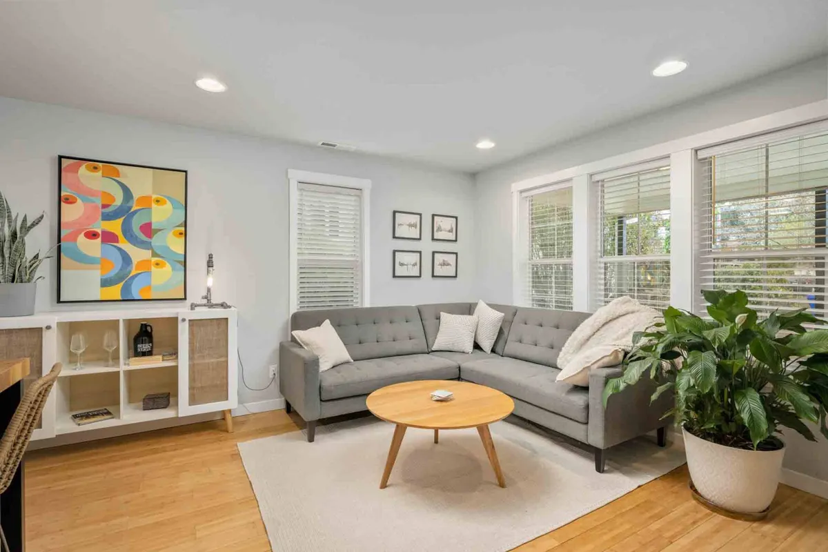 A cozy living area featuring a light gray sectional sofa, a wooden coffee table, a green corner plant, and vibrant abstract artwork. The setup encourages relaxation, socializing, or reading in a warm, peaceful atmosphere.