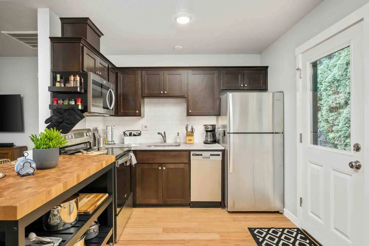 A warm, modern kitchen showcasing a large island, sleek stainless steel appliances, dark wooden cabinetry, and a bright white backsplash. The space is filled with natural light and provides an inviting environment for cooking, entertaining, and gathering.