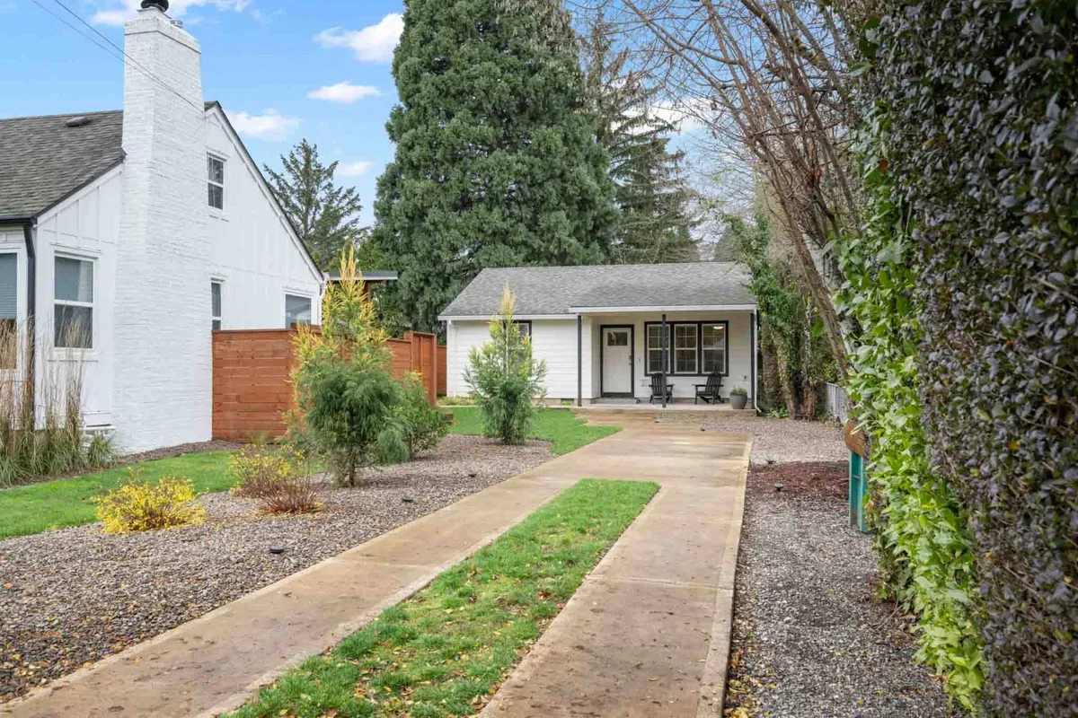 The exterior of the Bridge City Casita is showcased, featuring a cozy porch, neatly manicured garden, and a gravel driveway. The scene suggests a peaceful and inviting setting, ideal for relaxing outdoors or enjoying quiet mornings before heading out for business or hospital visits.
