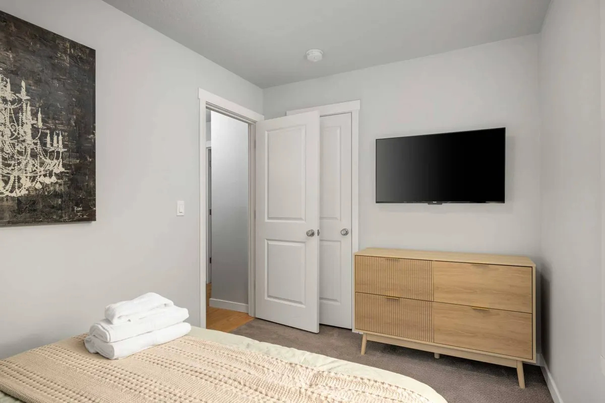 The image depicts a serene bedroom with a neatly made bed topped with rolled towels, a television, a textured dresser, and subtle artwork. The space exudes a sense of personal retreat and comfort, ideal for unwinding after a long day.
