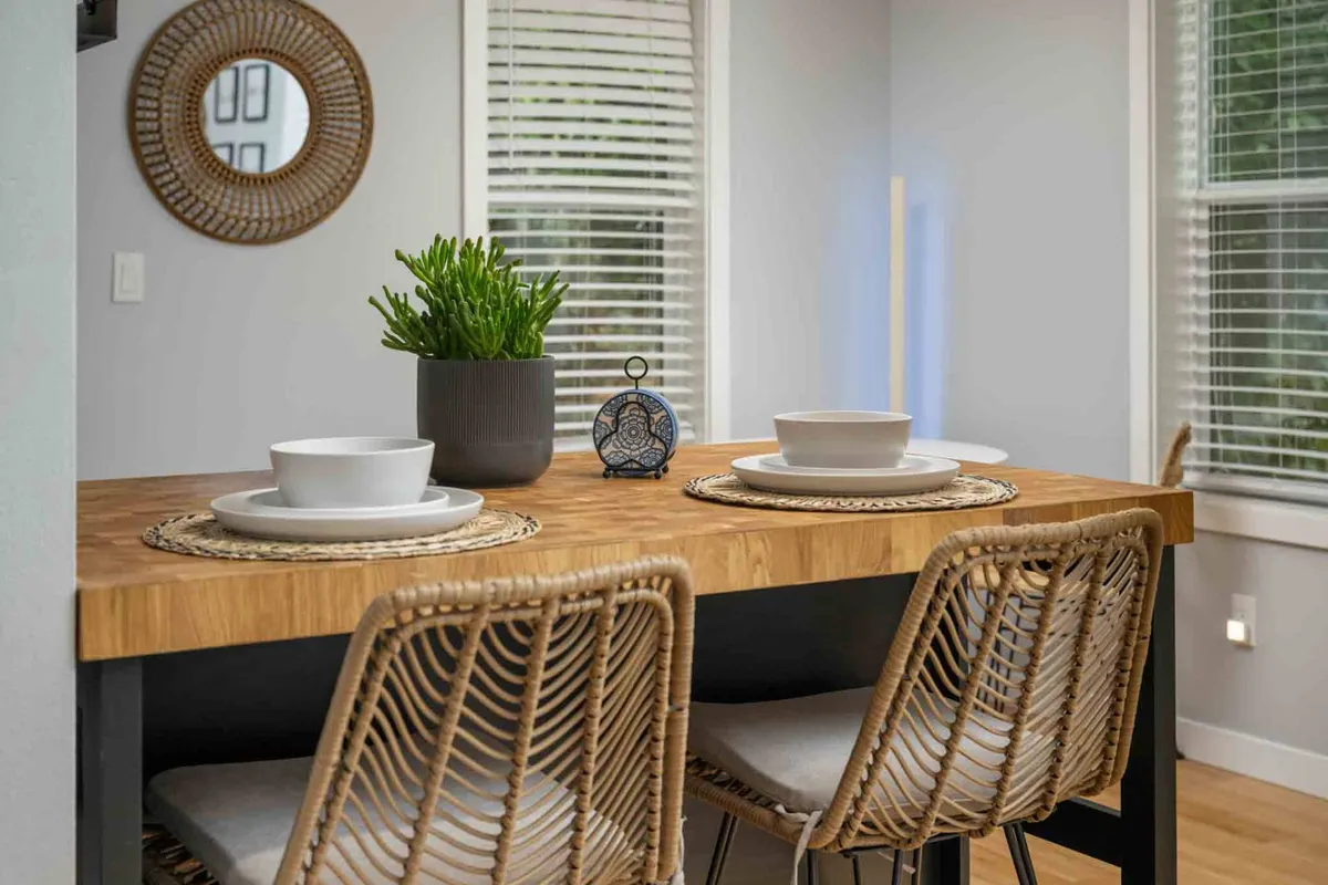 A cozy, sunlit dining nook featuring a wooden table set with dinnerware and woven placemats, a touch of greenery from a potted plant, and a mirror that enhances the sense of space. The setup suggests an inviting spot for relaxed meals or morning coffee.