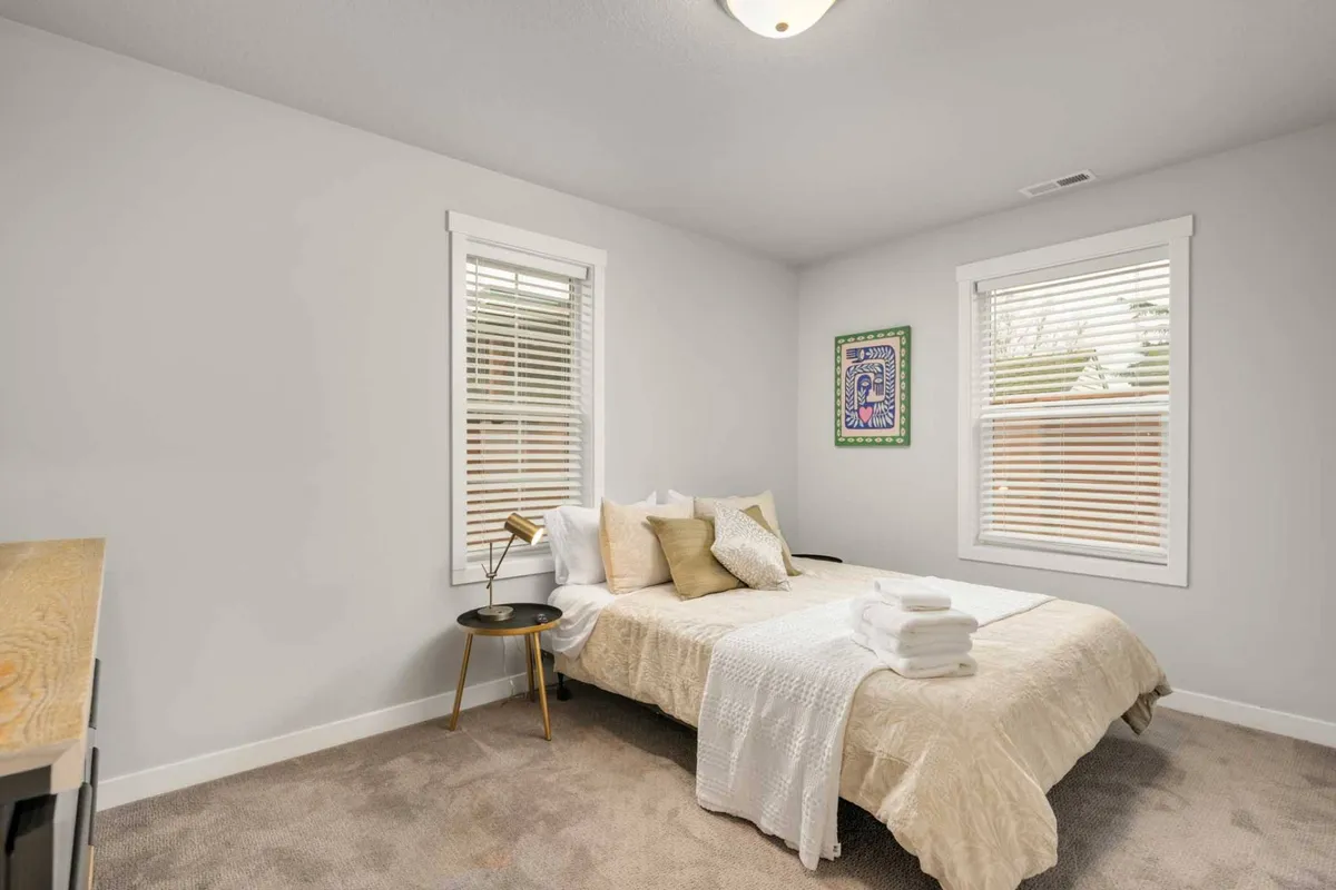 A tranquil bedroom with a cozy bed adorned with decorative pillows, modern artwork on the wall, and a stylish table lamp on the side table. Sunlight filters softly into the space, contributing to a peaceful, welcoming vibe ideal for quiet reflection or deep rest.