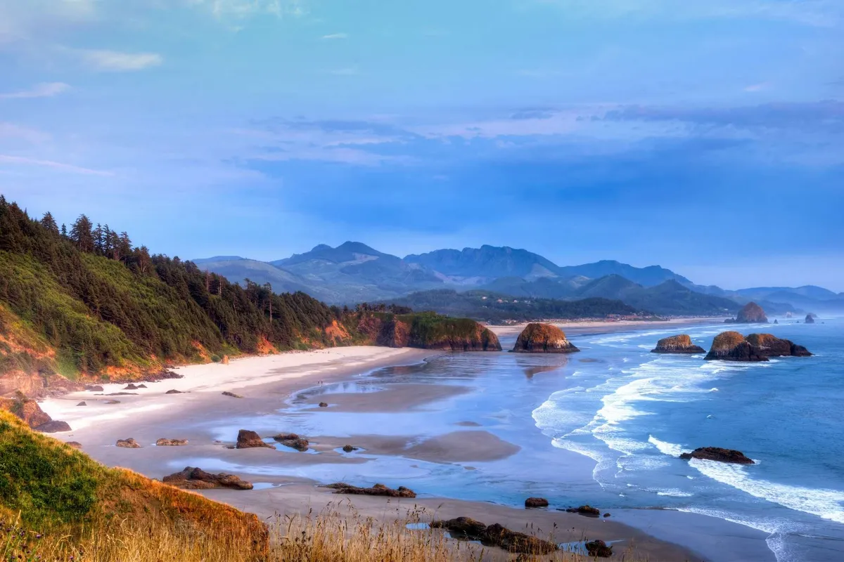 A scenic view of the Oregon coast with rolling ocean waves, dramatic rock formations, sandy beach, and lush forested mountains in the distance. The tranquil setting evokes outdoor adventure and natural exploration.
