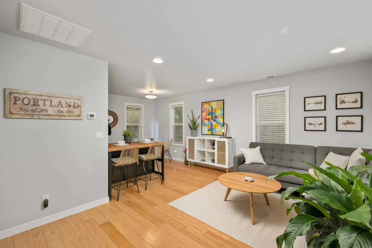 The image showcases a cozy, light-filled living and dining area featuring a stylish dining table set for sharing meals, a comfortable sofa, lush green plants, and curated artwork—including a nod to Portland’s vibrant spirit. The overall ambiance is warm and welcoming, inviting guests to relax, socialize, and appreciate the artistic touches.