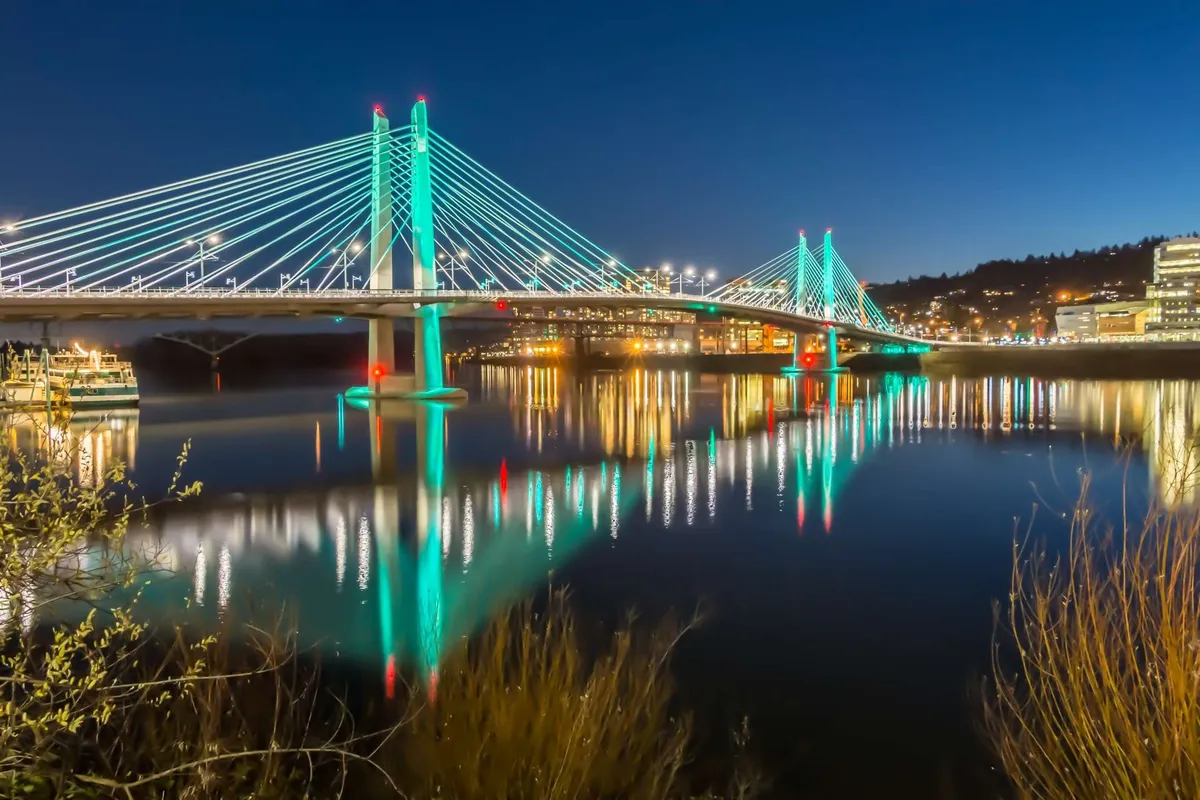 A vibrant view of the city skyline across the river at dusk, with a lit bridge and reflections shimmering in the water. The scene captures the proximity to urban life, city events, and the energy of Portland's downtown.