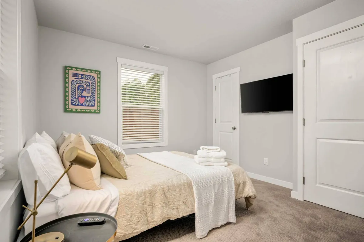 This image features a bedroom with a cozy bed adorned with soft linens, tasteful artwork on the wall, a television, and a window with blinds subtly filtering the natural light. The entire scene conveys an inviting, tranquil setting where one can easily unwind, read, or catch up on rest.