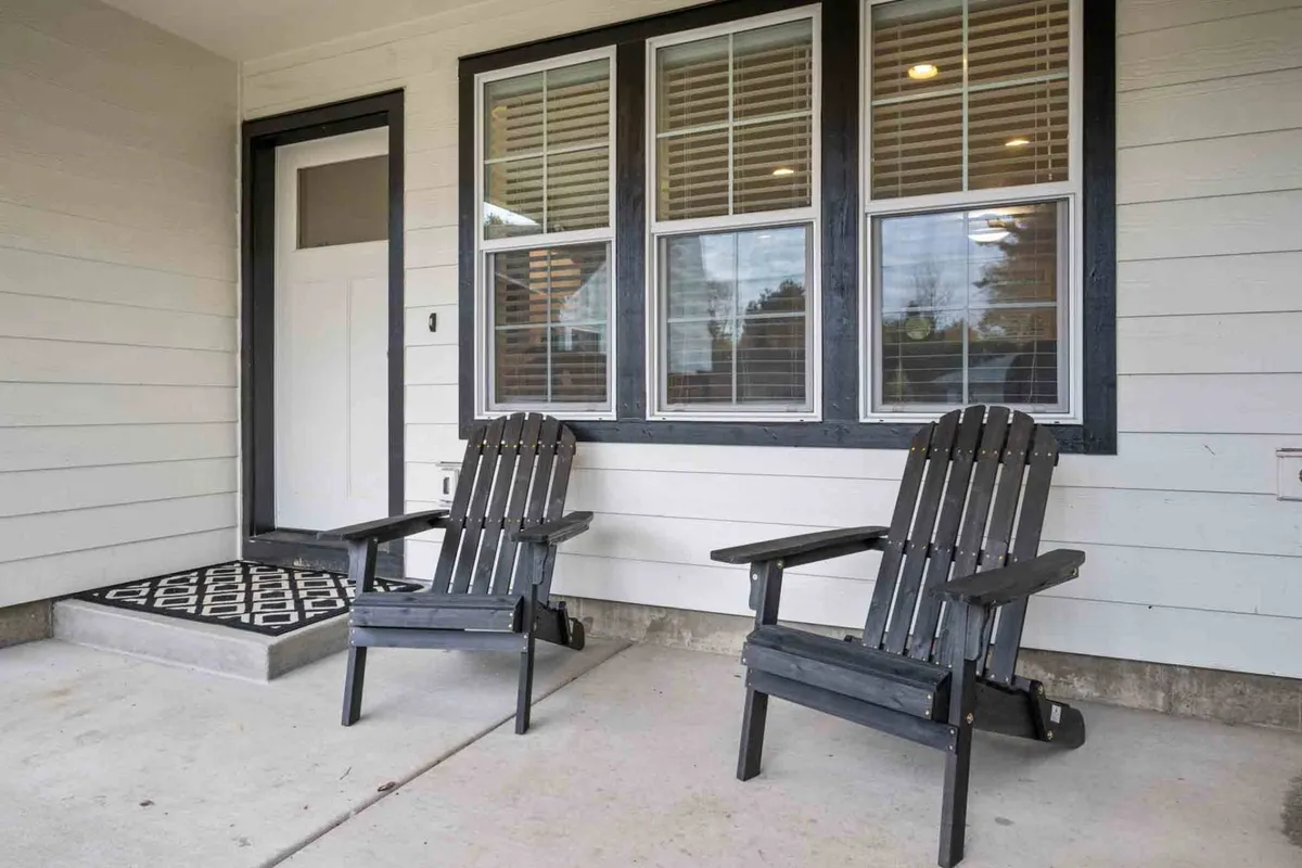 This exterior shot highlights a front porch with black Adirondack chairs, a decorative welcome mat, and expansive windows. The scene bathes in morning sunlight, evoking a sense of calm and inviting guests to enjoy quiet moments outside.