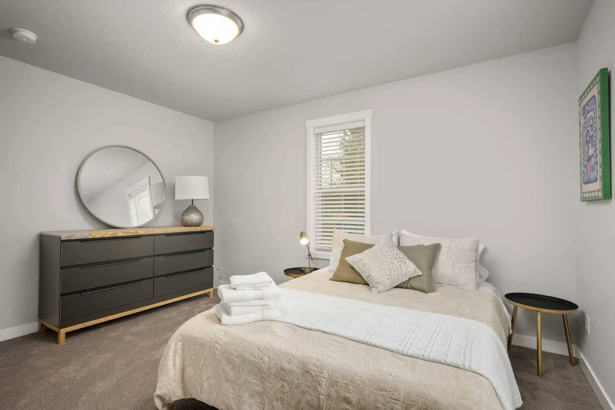 A serene bedroom featuring a plush queen bed dressed with soft, decorative pillows and luxurious linens. A round mirror above a sleek dresser, a modern nightstand with a warm lamp, and gentle lighting create a peaceful, inviting atmosphere. The overall color palette is soft and calming, perfect for unwinding after a long day.